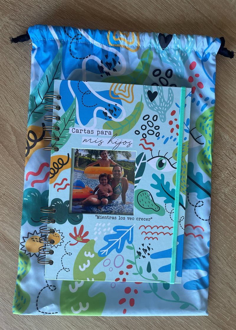 Cuaderno Cartas para mis hijos: agenda argollada de tapa azul claro con ilustraciones de hojas y soles, ventana con una foto familiar, elástico menta y su bolsa de tela a juego.